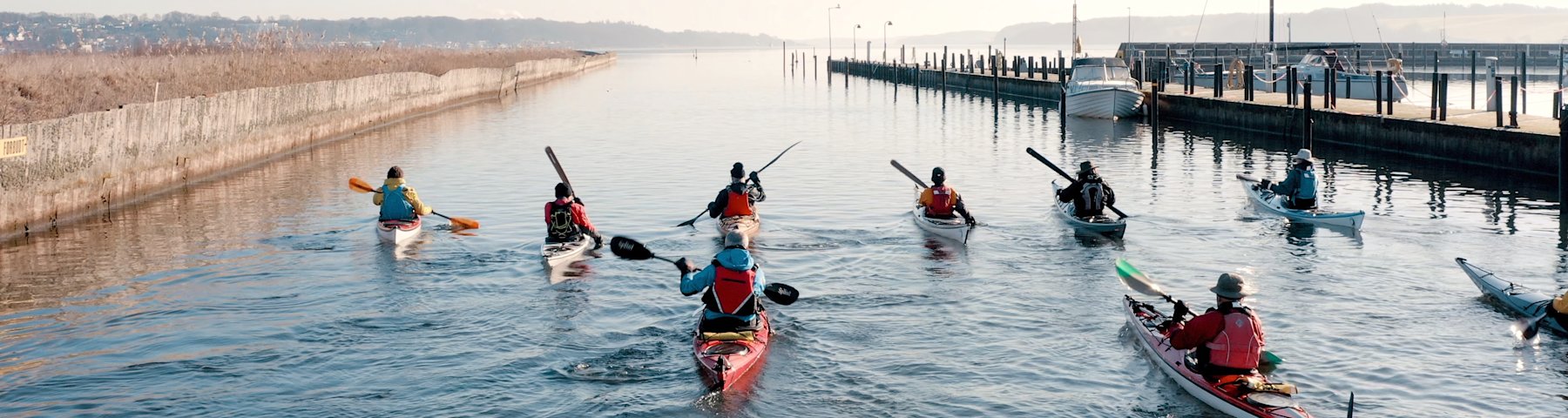 En flok roere i kajakker sejler ud mod Kolding Fjord fra Kolding Havn. De sejler forbi lystbådehavnen, som er i højre side af billedet