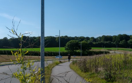 Ny udstykning af byggegrunde ved Sønderkobbel i Christiansfeld i 2021. Udstykningen ligger som en grøn mark i baggrunden. Forrest og midt i billedet snor en vej og en cykelsti sig ved siden ad hinanden frem mod udstykningen og et skovbryn, der afgrænser udstykningen bagest i billedet. Der går en person på cykelstien.