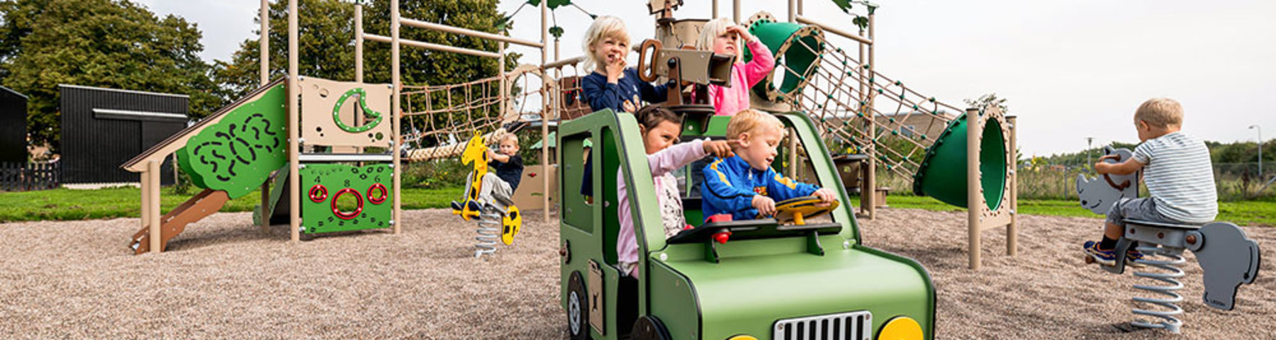 Legeplads med safaritema med Landrover i forgrunden, hvor fire børn leger - til højre i billedet sidder et barn på en af legepladsens vippedyr - en hest