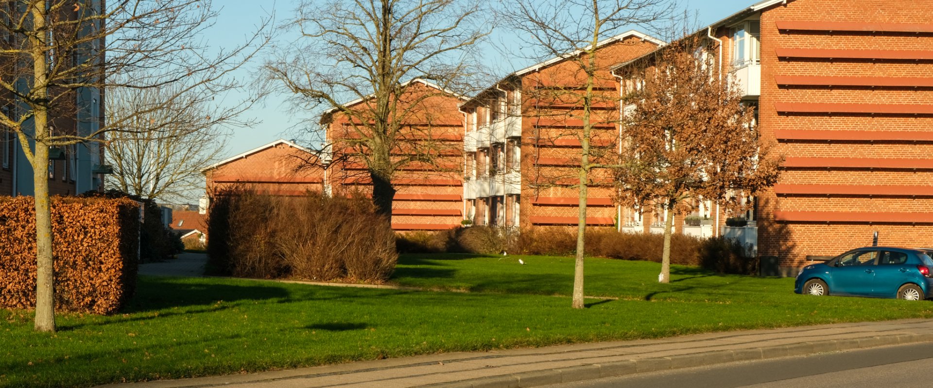 En stribe på 4 røde boligblokke med hvide altaner i Brændkjær-kvarteret i Kolding med grøn græsplæne, træer og hække