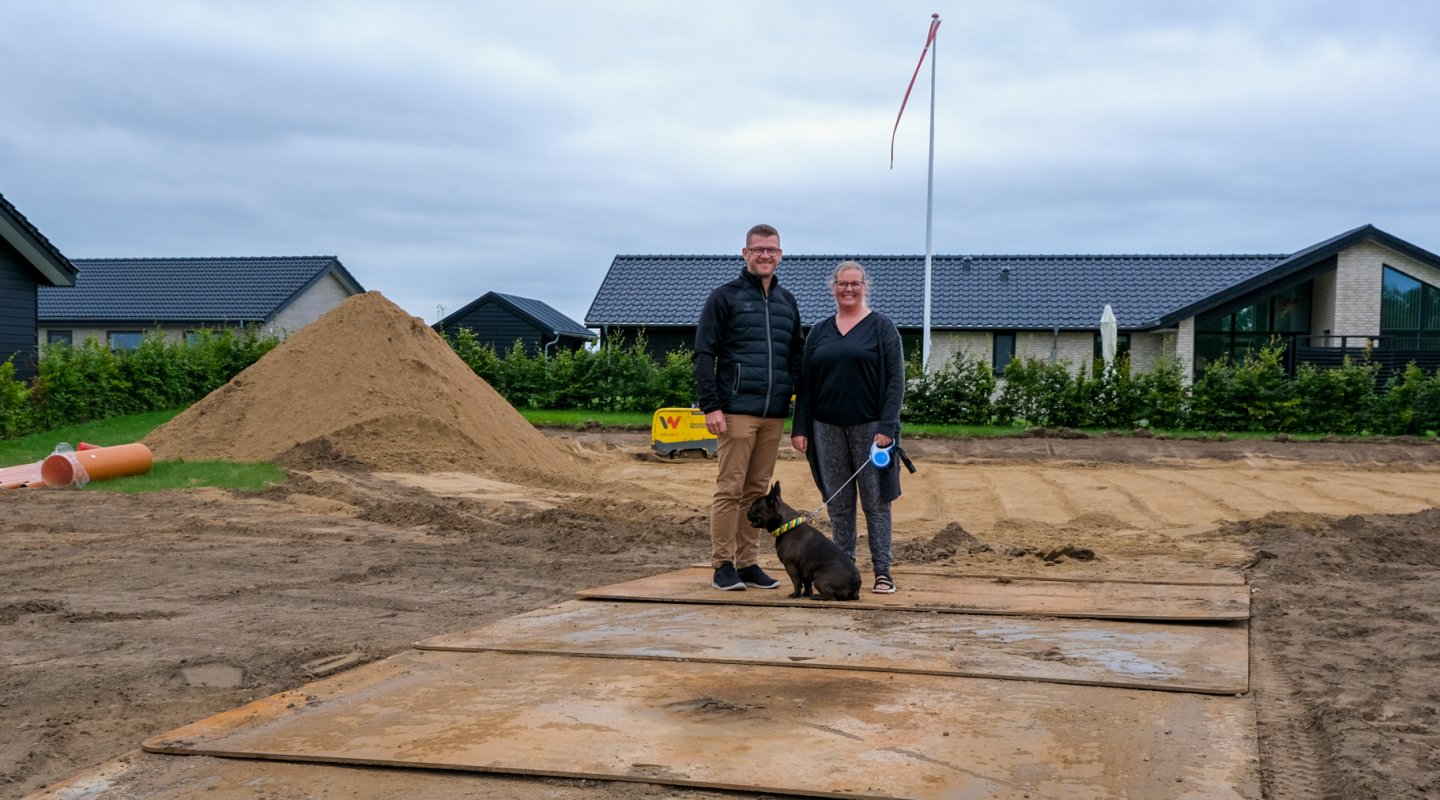 Thomas og Christina Graugaard-Nielsen står midt på deres byggegrund på Vesterengen i Viuf. Thomas står til venstre, Christina står til højre. Foran dem ligger deres hund Svend. Der er  bunker af jord på grunden