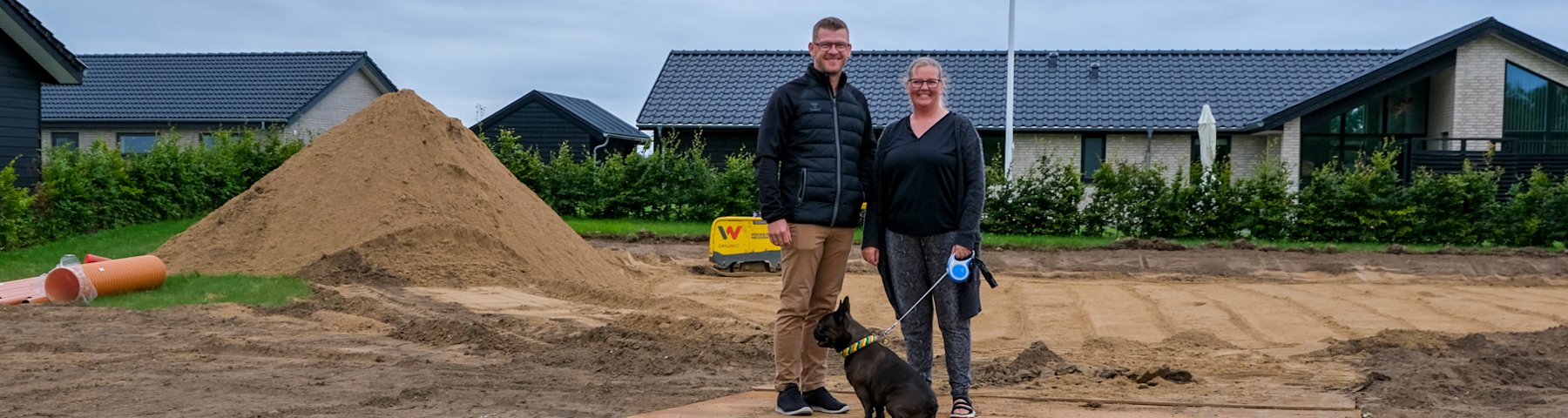 Thomas og Christina Graugaard-Nielsen står midt på deres byggegrund på Vesterengen i Viuf. Thomas står til venstre, Christina står til højre. Foran dem ligger deres hund Svend. Der er  bunker af jord på grunden
