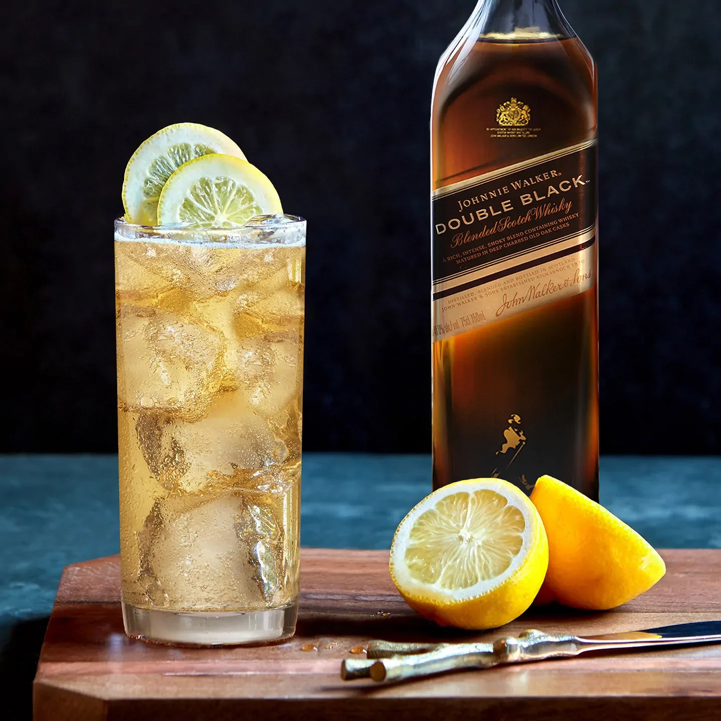 A glass of whiskey and ice with lemon wedges next to a bottle of Johnnie Walker Double Black blended scotch whisky on a wooden cutting board. 