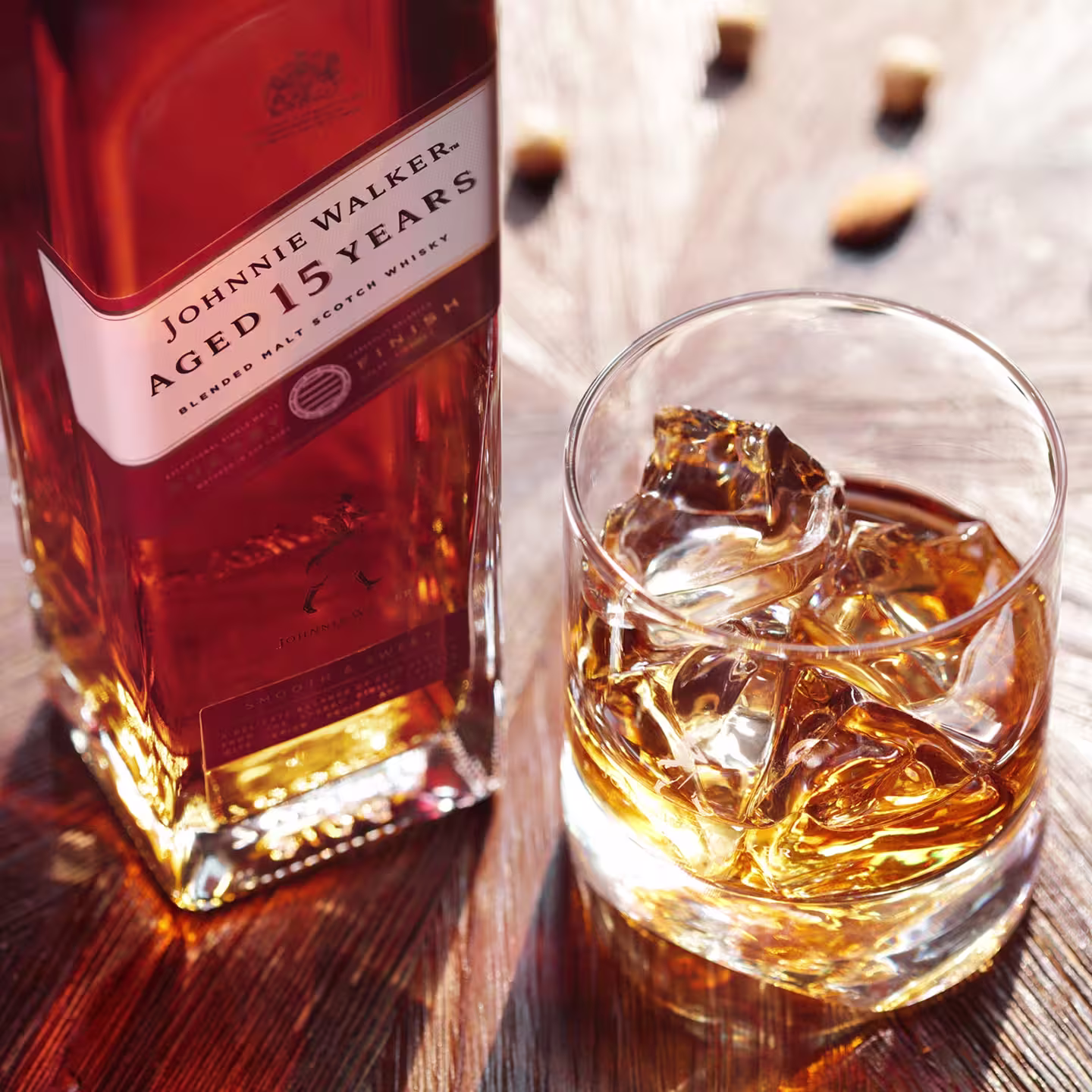 A glass of whisky with ice sits next to a bottle of Johnnie Walker Aged 15 Years blended Scotch whisky on a wooden surface, with bottle caps blurred in the background.