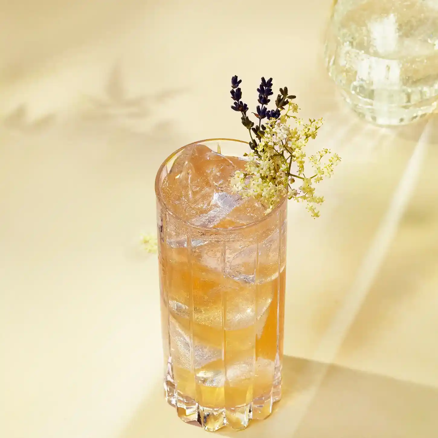 Un vaso alto lleno de una bebida de color ámbar claro, cubitos de hielo y adornado con pequeñas flores blancas y una ramita de lavanda, reposa sobre una superficie pálida bañada por una suave luz natural. Se ven sombras de flores en el fondo.