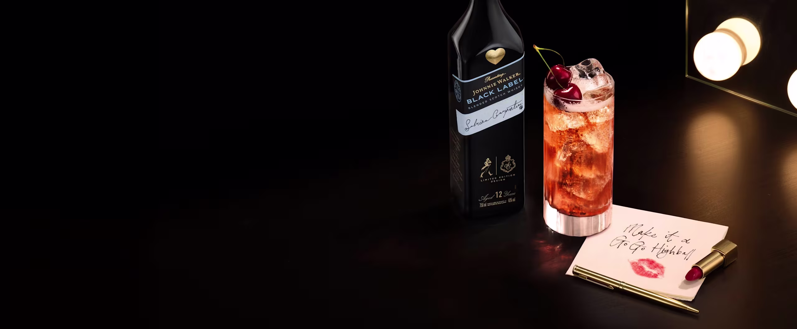 Tall cocktail with ice, cherry garnish, Johnnie Walker Black Label Sabrina Carpenter Limited Edition Design whisky bottle, lipstick, and note by lit mirror.