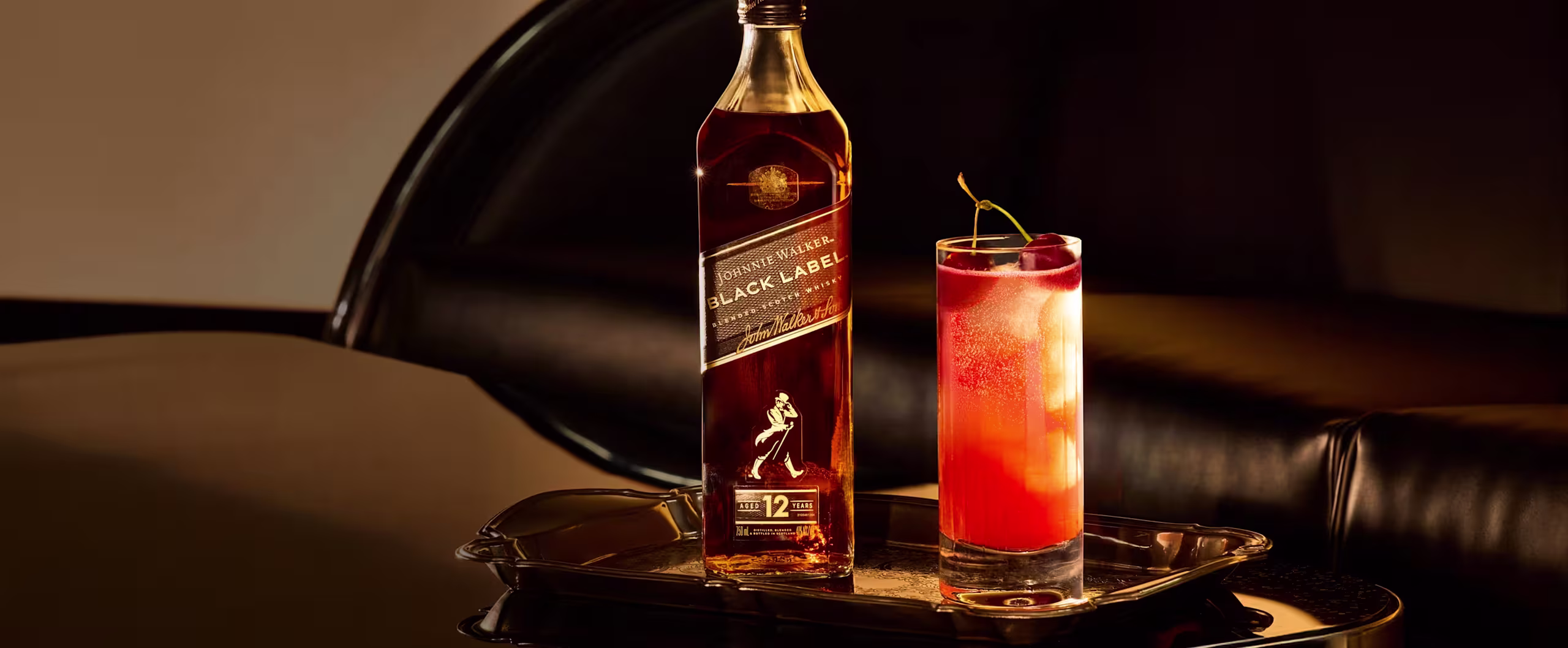 Een fles Johnnie Walker Black Label whisky en een grote rode cocktail, versierd met een vleugje citrus, staan ​​op een glanzend dienblad op een glanzend zwarte tafel. Op de achtergrond staat een zwarte leren bank.