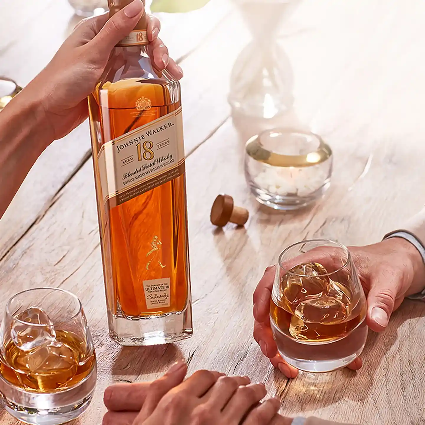 มือทั้งสองข้างถือแก้ววิสกี้บนโต๊ะไม้ พร้อมขวดวิสกี้ Johnnie Walker 18 Year Old และที่ใส่เทียนแก้วทรงกลม แสงแดดส่องลงมาเพื่อสร้างบรรยากาศที่อบอุ่นและน่าดึงดูด.