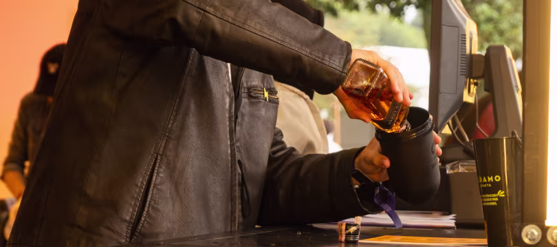 Una persona con una chaqueta de cuero negra vierte un líquido ámbar de una botella de whisky en una taza negra en un bar al aire libre o en el mostrador de un evento.