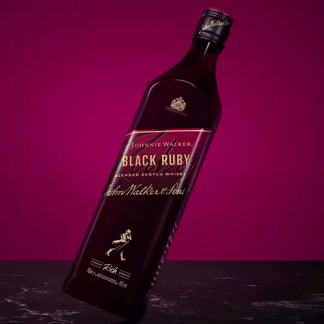  Nahaufnahme einer Flasche Johnnie Walker Black Ruby Blended Scotch Whisky vor dunkelrosa Hintergrund, zu sehen sind das Etikett mit goldenen und rosa Akzenten und das ikonische Johnnie Walker-Logo.