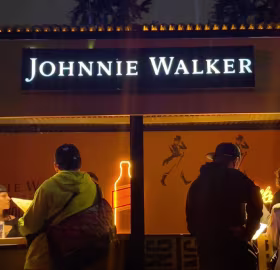 Una escena nocturna muestra un cartel de Johnnie Walker sobre un puesto. Tres personas están de pie frente al puesto, interactuando con el encargado. El contorno de neón de una botella y el logotipo de Johnnie Walker iluminan la zona.