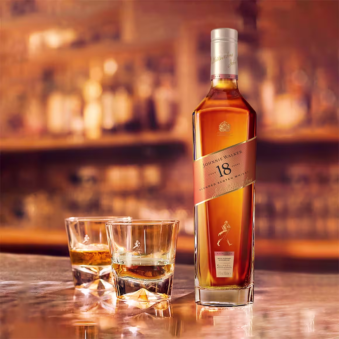 A bottle of Johnnie Walker 18-year-old blended Scotch whisky stands on a bar next to two glasses filled with whisky, with a softly blurred bar background.