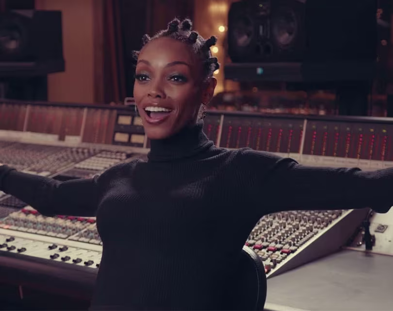 Uma mulher com cabelo trançado sorri de braços abertos, parada em frente a um grande console de mixagem de áudio em um estúdio de gravação. Ela veste um suéter preto de gola alta.