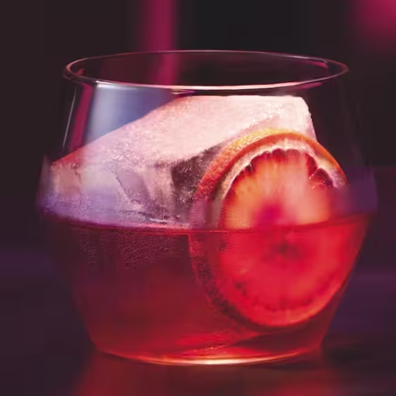 A glass of red cocktail with a large ice cube and an orange slice sits on a dark surface, with dramatic pink and purple lighting in the background.