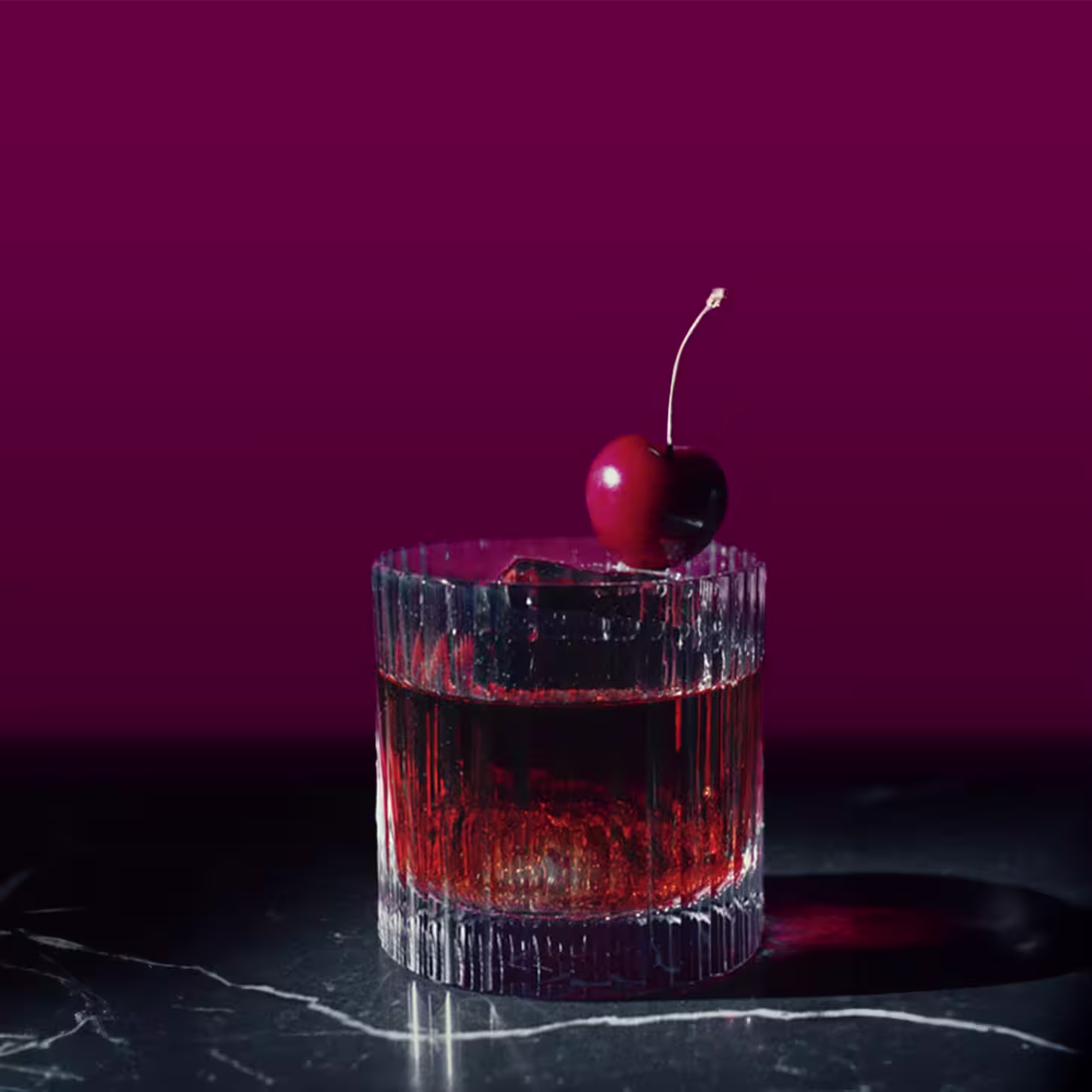 Una copa de cóctel de color rojo oscuro se asienta sobre una superficie de mármol sobre un fondo marrón oscuro. La bebida está adornada con una cereza en el borde.