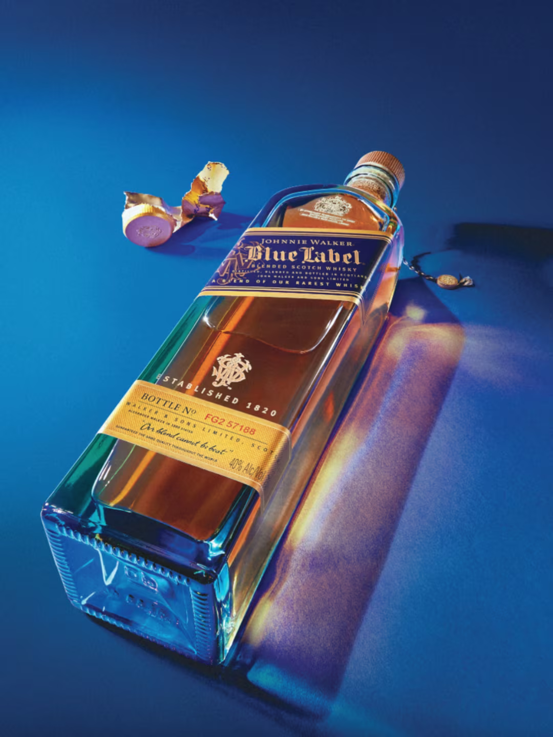 A bottle of Johnnie Walker Blue Label whisky lies on a blue surface with dramatic lighting. The gold cap is off and placed nearby, and the bottle casts a soft, colorful shadow.