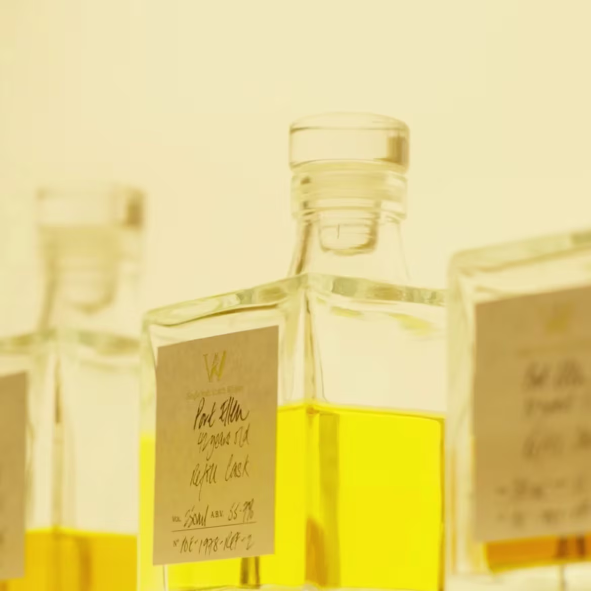 Square glass bottles filled with amber liquid are arranged in a soft, warm light. The bottles have handwritten labels featuring decorative typography.