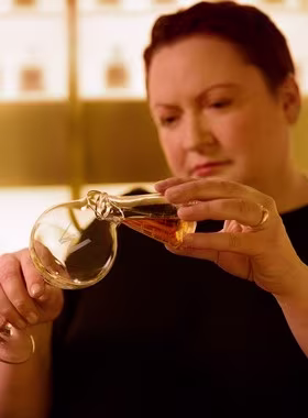 A person with short hair pours liquid from a round-bottom flask into a smaller vessel, focusing intently on the task. The background is softly lit and blurred.