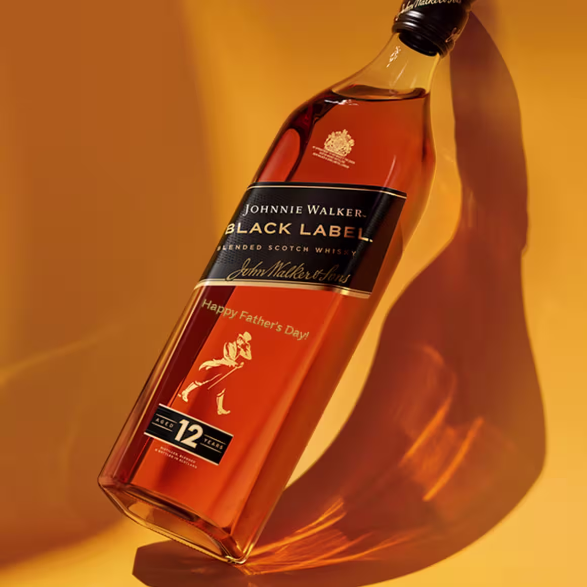 A Johnnie Walker Black Label 12-year-old blended Scotch whisky bottle lies on its side against a golden orange background, casting a soft shadow. The label reads Happy Fathers Day.