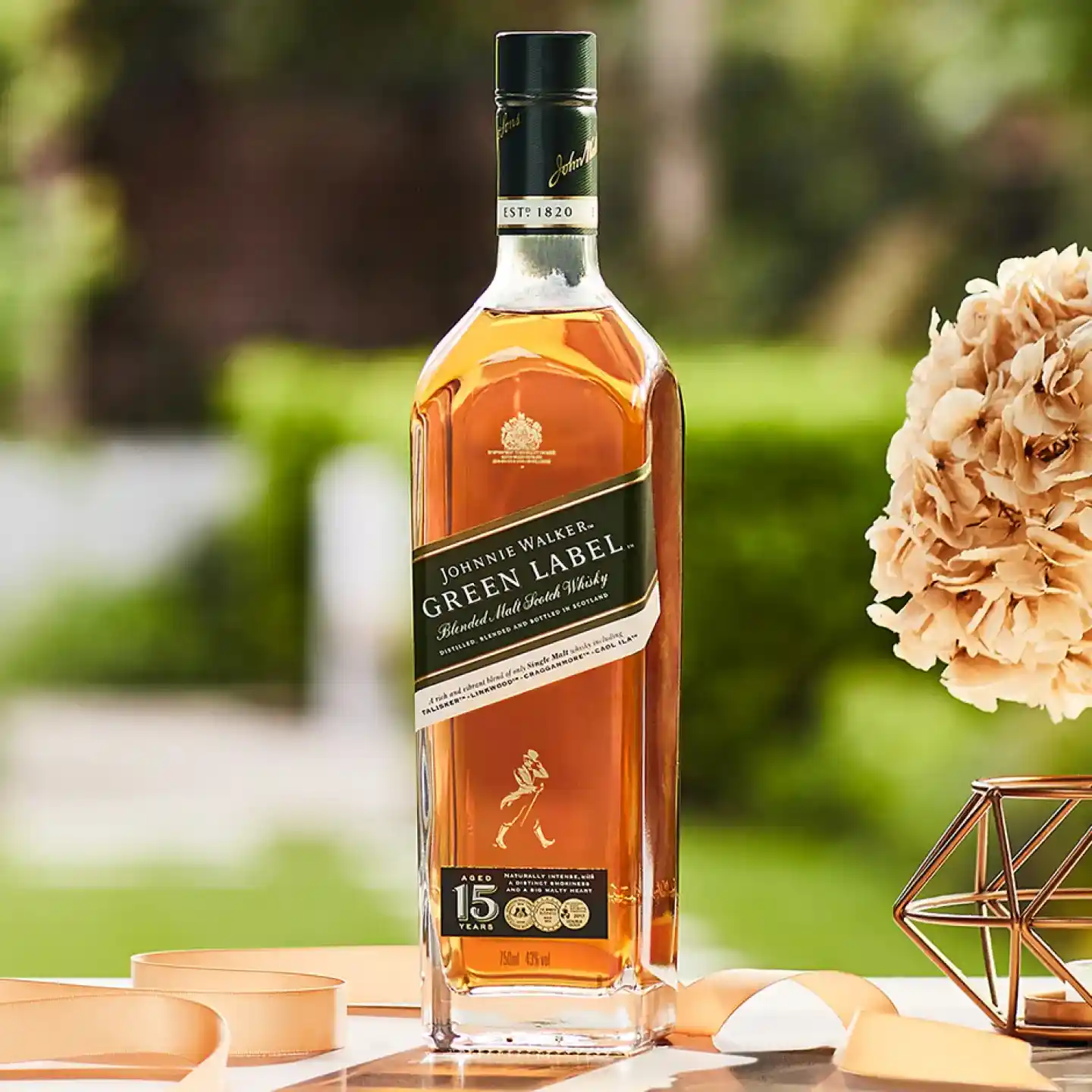 Een fles Johnnie Walker Green Label Blended Malt Scotch Whisky staat buiten op een tafel. Er staat een boeket beige bloemen rechts en een lint is gedeeltelijk zichtbaar op de tafel. De achtergrond is wazig groen.