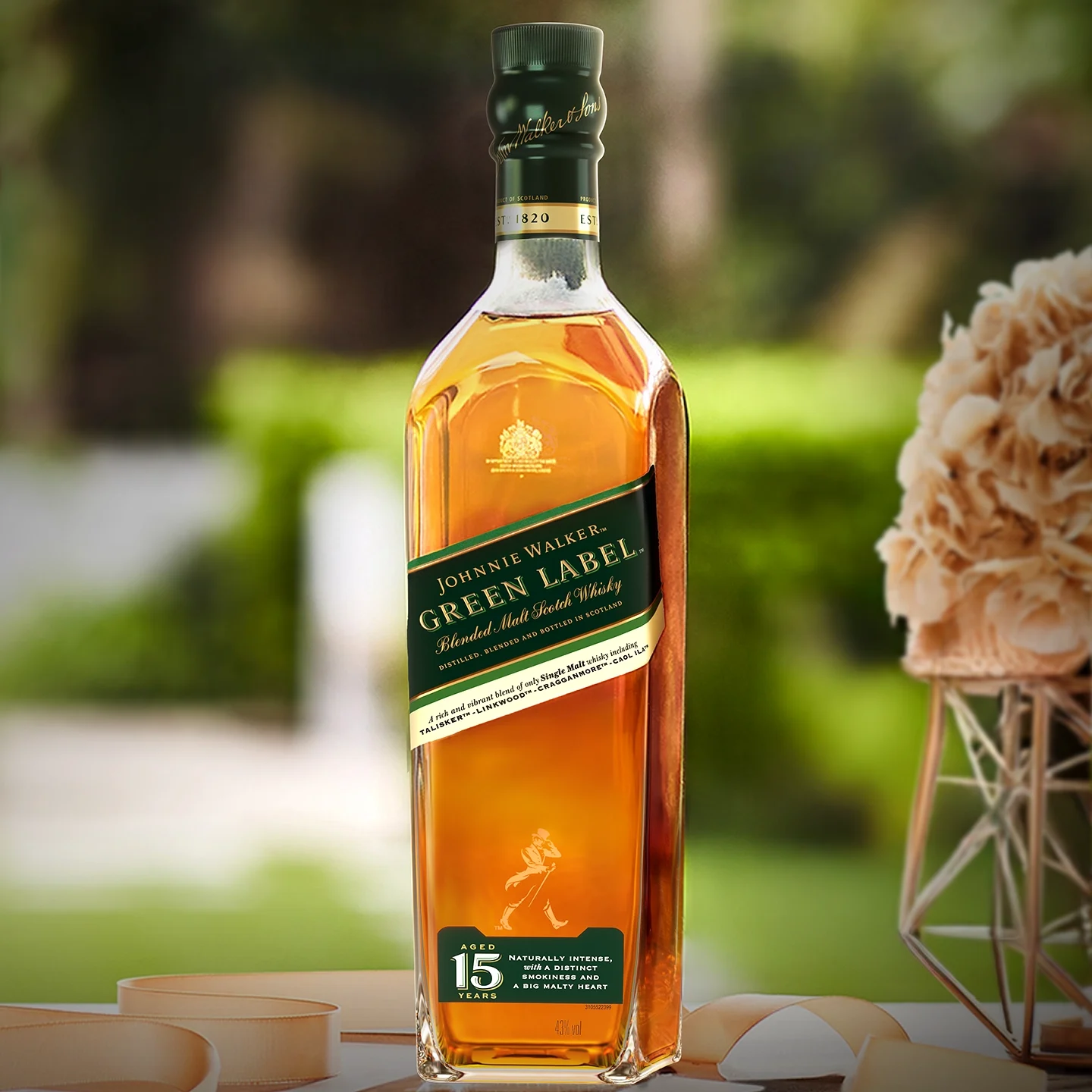 Una botella de whisky escocés de malta mezclado Johnnie Walker Green Label de 15 años se encuentra sobre una mesa al aire libre, con un fondo verde borroso y elementos decorativos cerca.