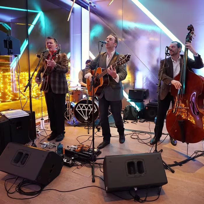 Four musicians perform indoors with guitars, drum set, and upright bass, surrounded by colorful lights.