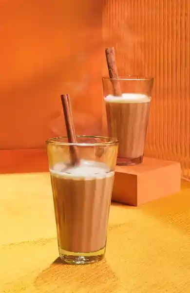 Two glasses of steaming brown liquid with cinnamon sticks sit on a yellow and orange surface. 
