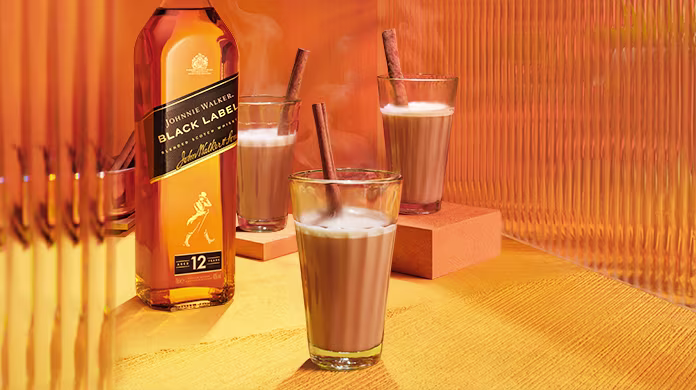 Una botella de whisky Johnnie Walker Black Label se encuentra junto a tres vasos de cremosos cócteles, cada uno adornado con una ramita de canela, sobre una mesa de tonos naranja brillantes con paneles texturizados al fondo.