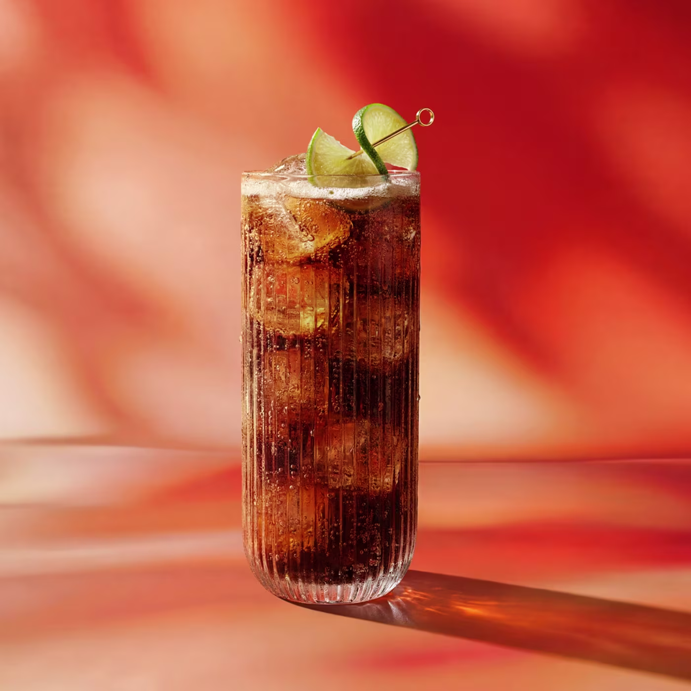 A tall glass of cola with ice and lime garnish sits on a warm, orange-red background.