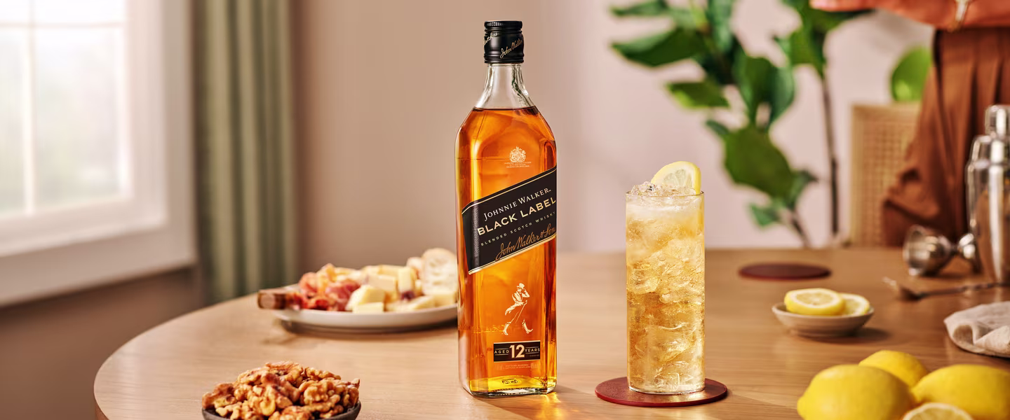 A bottle of Johnnie Walker Black Label whisky stands on a table beside a tall glass of whisky with ice and a lemon slice. The table also has walnuts, cheese, and lemon halves, with a blurred background.