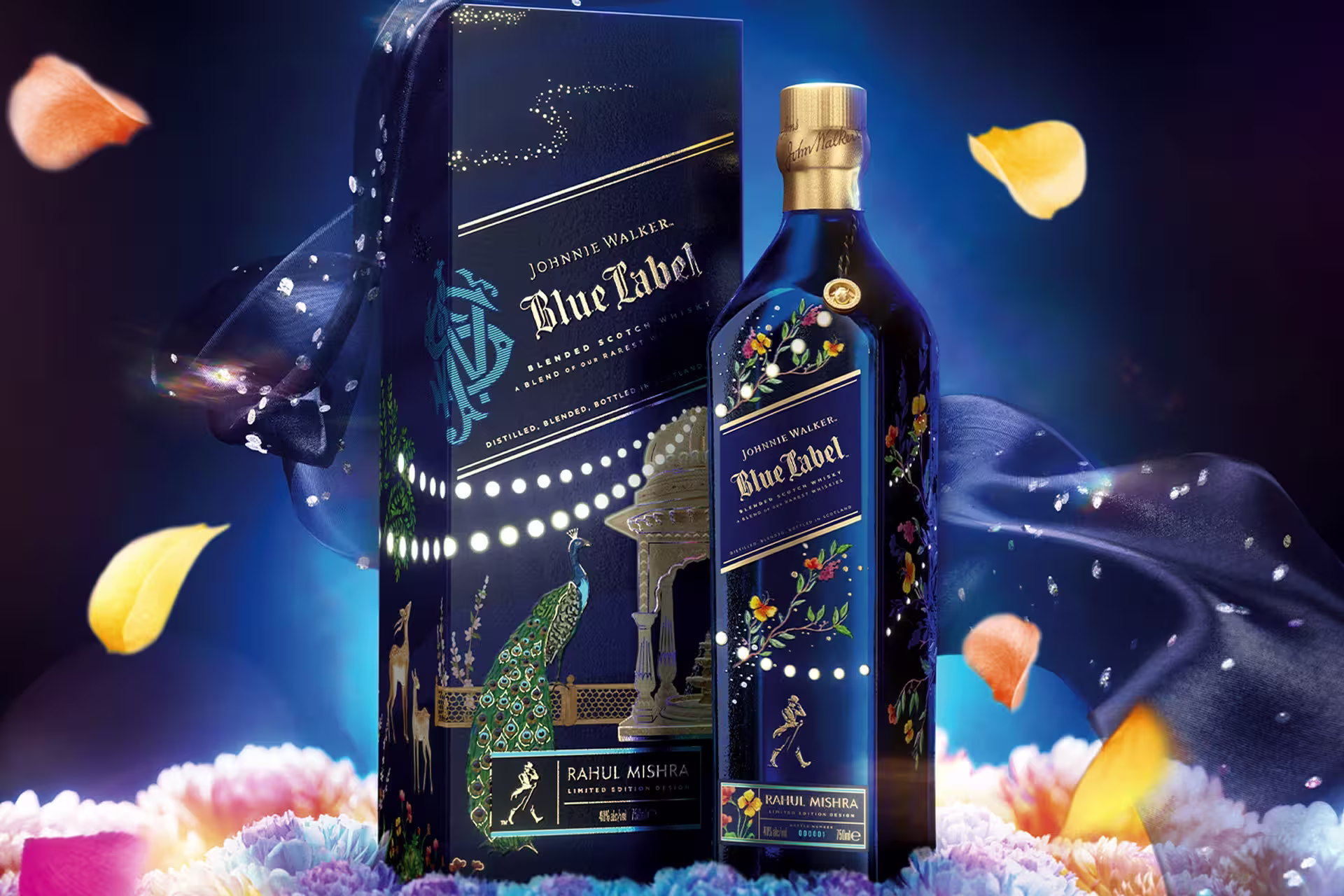 A limited edition Johnnie Walker Blue Label bottle and box, designed by Rahul Mishra, featuring intricate, colourful patterns with gold accents and floral motifs, surrounded by flower petals and a decorative silky fabric.