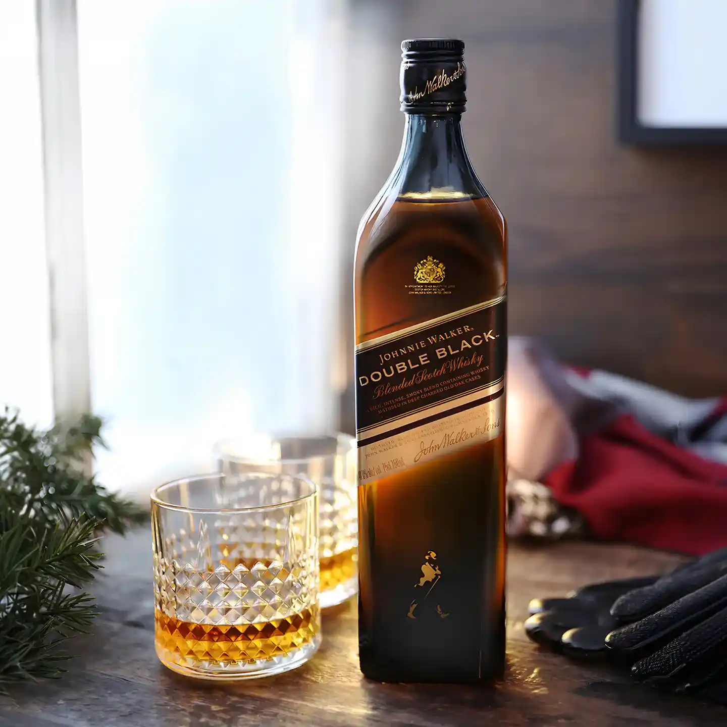 Johnnie Walker Double Black 위스키 한 병이 위스키로 가득 찬 잔 옆 나무 표면 위에 놓여 있습니다. 배경에는 창문, 약간의 녹색 식물, 부드러운 조명이 있어 아늑한 분위기를 연출합니다.