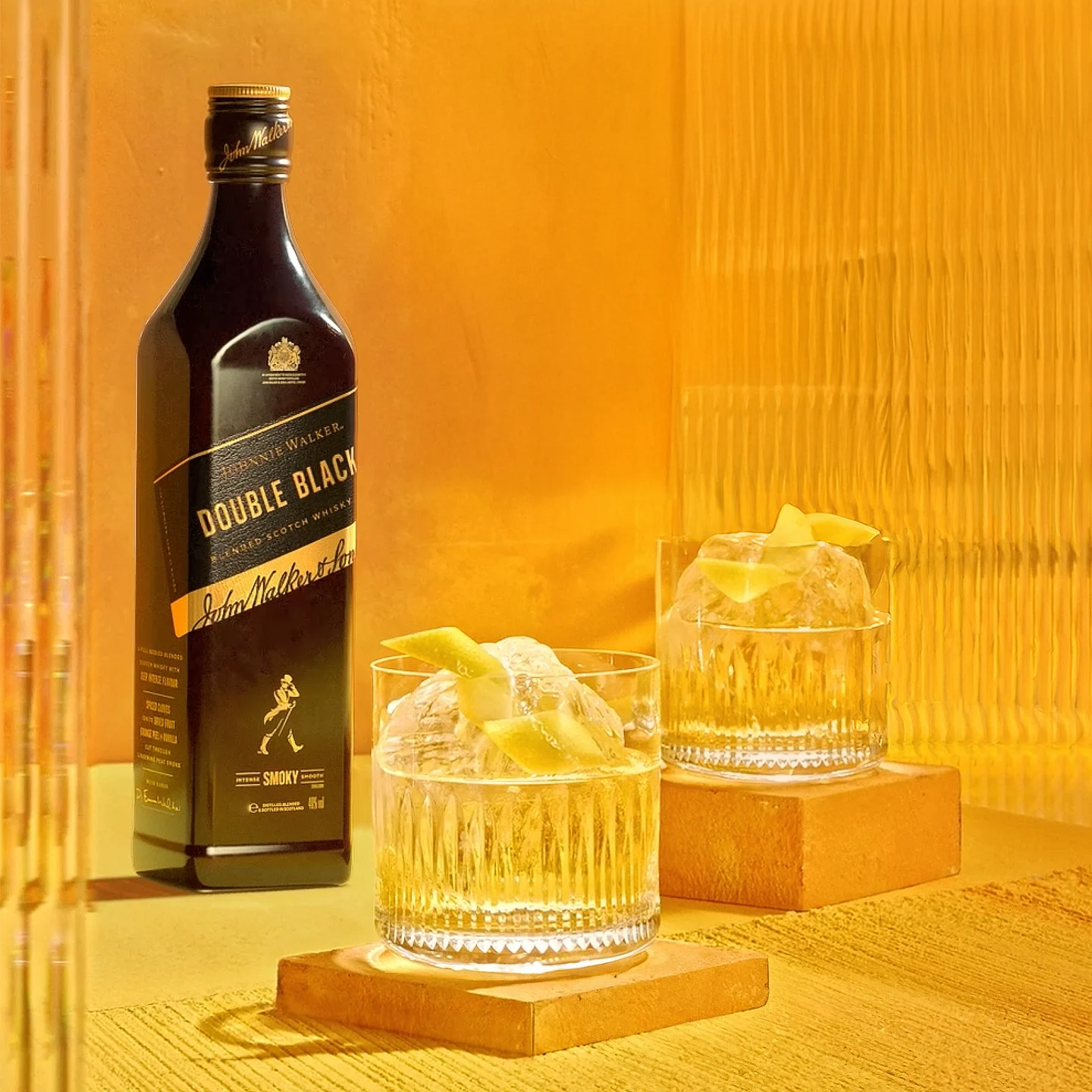 A bottle of Johnnie Walker Double Black whisky stands next to two glasses of whisky with large ice cubes and lemon twists, all set on golden-yellow blocks against a warm, amber-toned background.