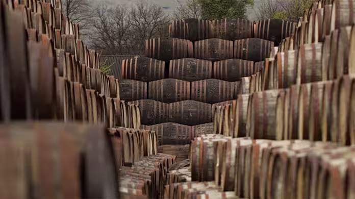  De grandes piles de tonneaux en bois sont disposées en rangées à l'extérieur, formant un étroit chemin entre eux. Des arbres nus et un ciel nuageux sont visibles en arrière-plan, suggérant une saison fraîche ou automnale.