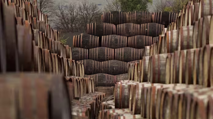 Large stacks of wooden barrels are arranged in rows outdoors, forming a narrow pathway between them. Bare trees and cloudy skies are visible in the background, suggesting a cool or autumn season.