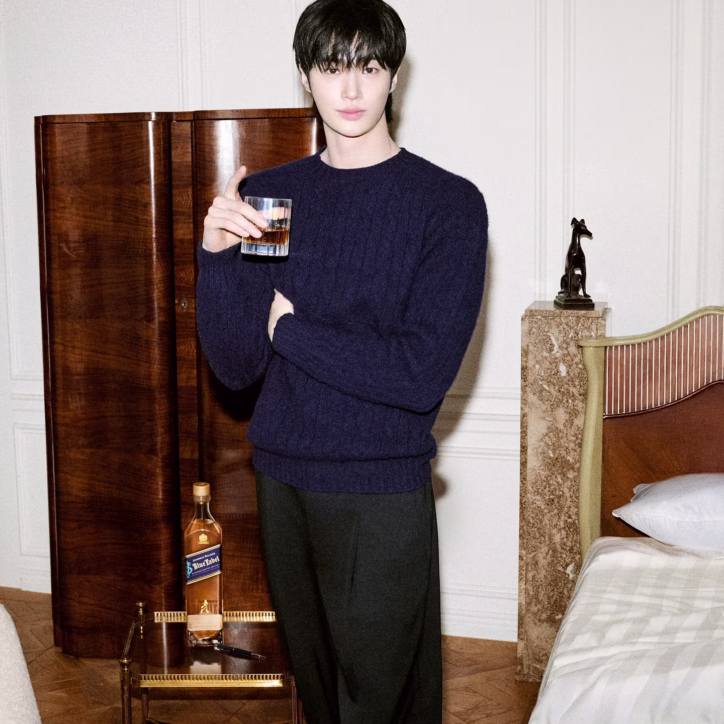 Person with short dark hair, wearing a navy sweater, holding a glass of whiskey, stands by a bed and nightstand.