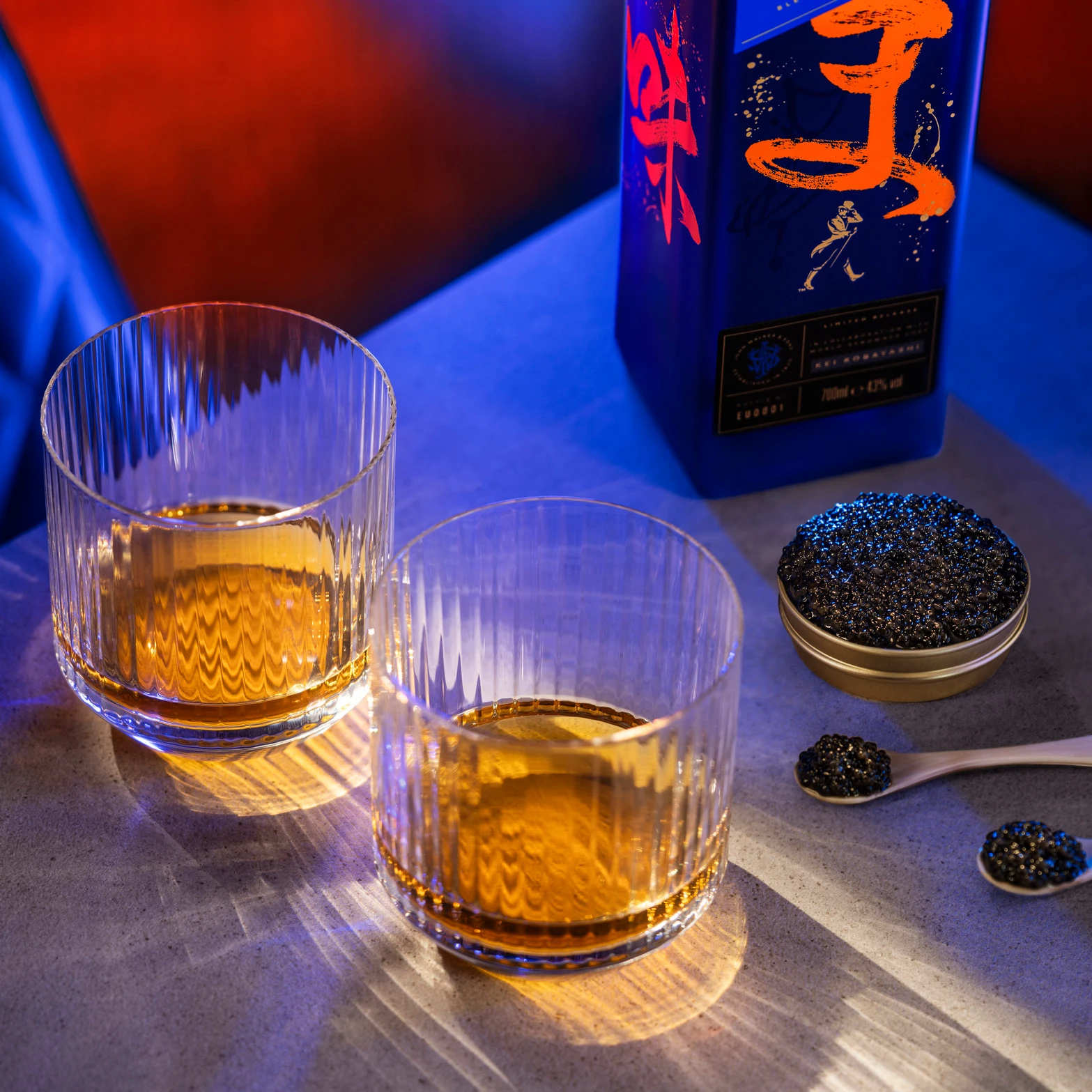 Two glasses filled with whisky on a table next to a Johnnie Walker Blue Label Elusive Umami bottle. A bowl of black caviar and a teaspoon are nearby. The setting is illuminated with blue and red lighting, creating a luxurious atmosphere.