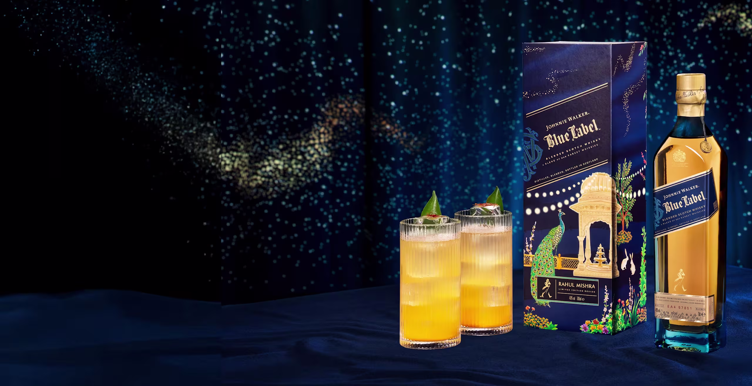 A bottle of Johnnie Walker Blue Label, its decorative box, and two orange cocktails with ice and green garnishes sit on a dark blue surface against a starry, dark background.