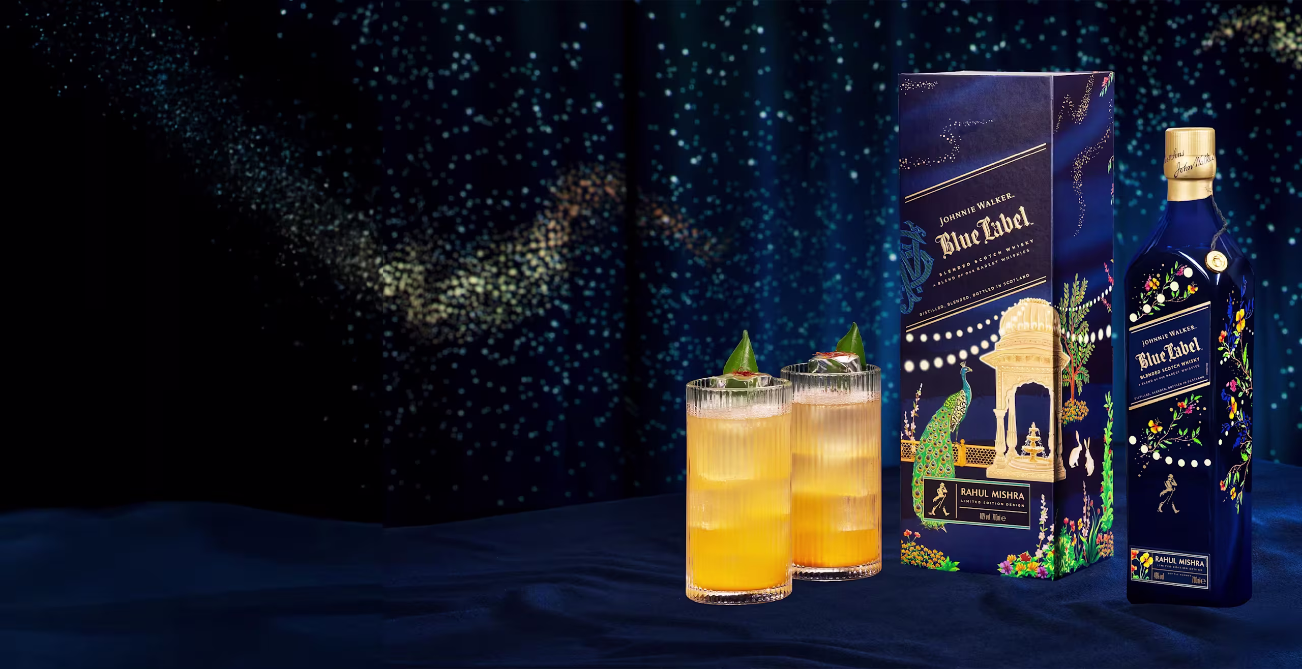A dark blue bottle of Johnnie Walker Blue Label next to its decorative box and two orange cocktails, all placed on a dark blue fabric with a starry, cosmic background.