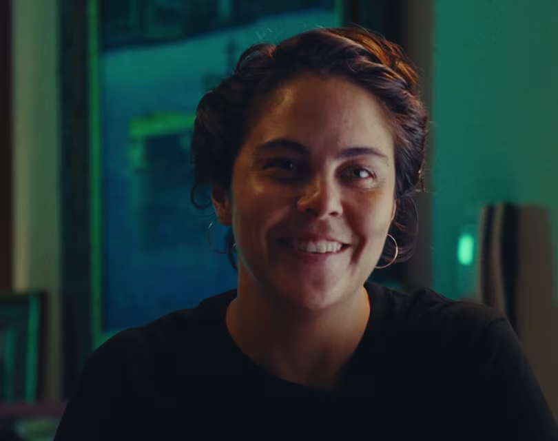 Una persona de pelo corto y oscuro, con una sonrisa amable, lleva pendientes de aro y una camisa oscura. Está sentada en un interior y el fondo está suavemente difuminado con toques de azul y verde.