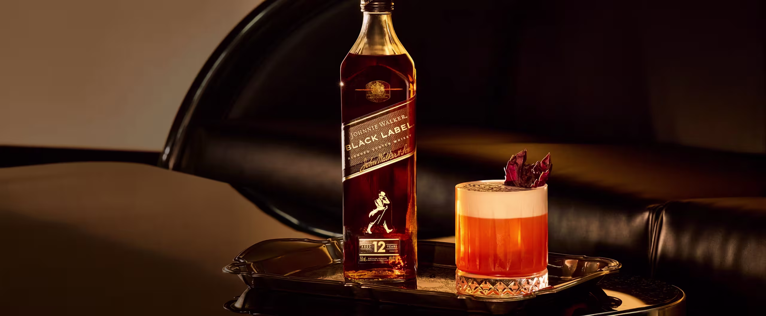Uma garrafa de uísque Johnnie Walker Black Label está ao lado de um coquetel decorado com flores secas, ambos dispostos em uma bandeja preta refletora sobre uma superfície brilhante com fundo escuro.