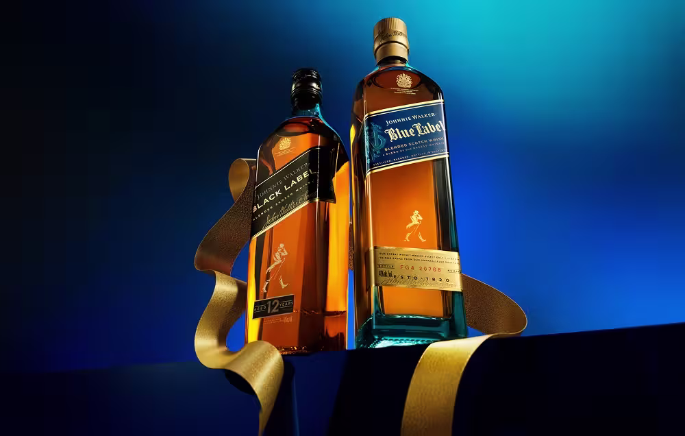 Zwei Flaschen Johnnie Walker Whisky – Black Label und Blue Label – stehen nebeneinander auf einer dunklen Oberfläche mit einem goldenen Band vor einem blauen Farbverlauf.