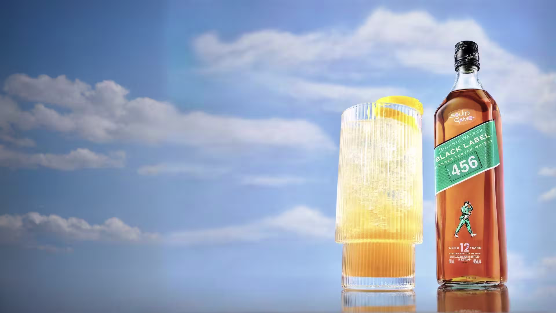 一瓶尊尼獲加黑牌456威士忌擺放在高腳杯旁，杯中盛著淺色飲品，旁邊放著一片檸檬。背景是湛藍的天空，點綴著朵朵白雲。