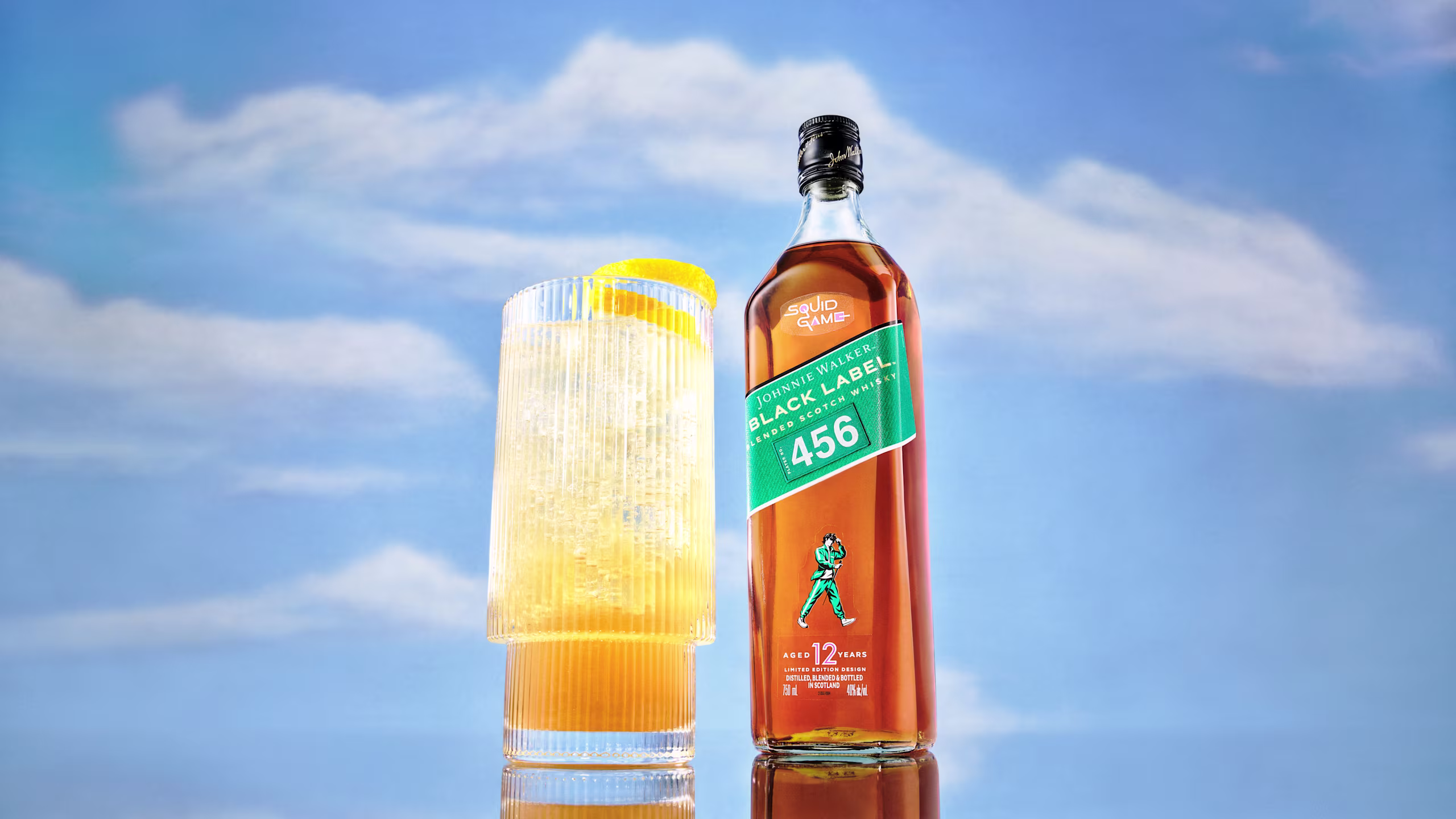 Een fles Johnnie Walker Black Label 456 whisky staat naast een groot glas met een lichtgekleurd drankje en een schijfje citroen. De achtergrond toont een helderblauwe lucht met donzige wolken.