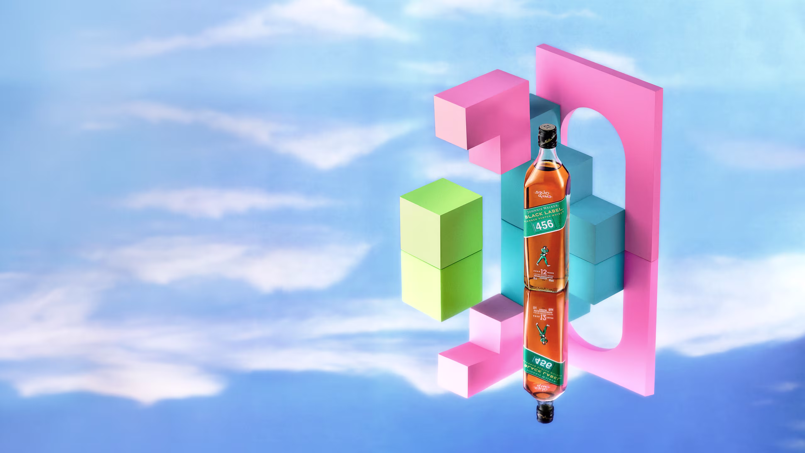 A bottle of Johnnie Walker Blue Label stands upright among colorful geometric blocks against a blue sky with clouds backdrop, with its reflection visible below.