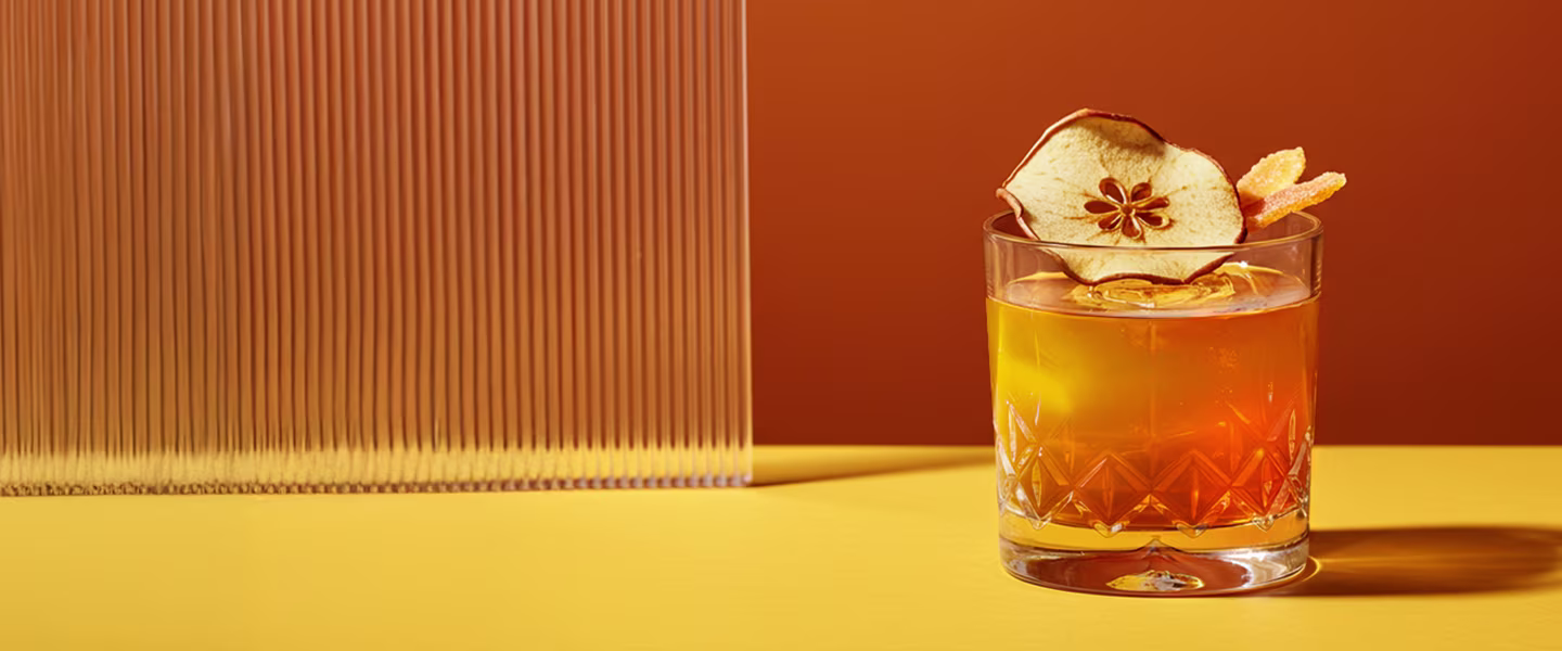 A hand holds a cocktail with a dried apple slice garnish on a marble table, next to a blue notebook, domino pieces, and part of another drink.