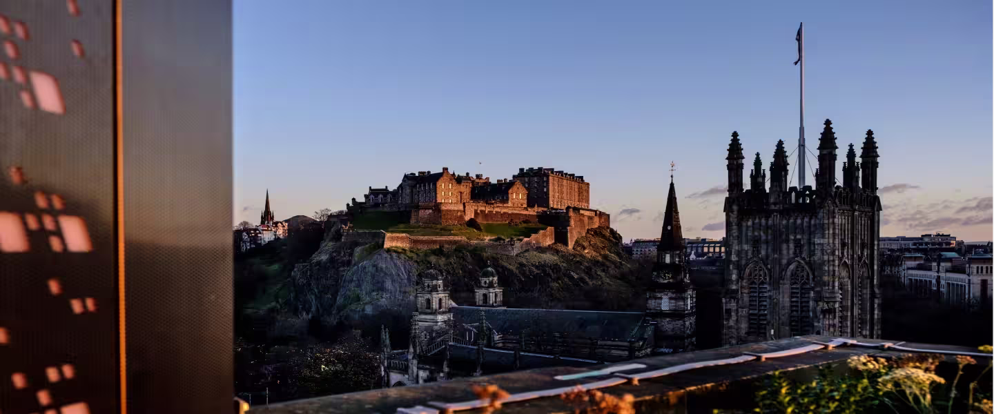 A panoramic view of Edinburgh Castle perched atop a volcanic crag, overlooking the city skyline. The castle stands majestically against a clear blue sky.