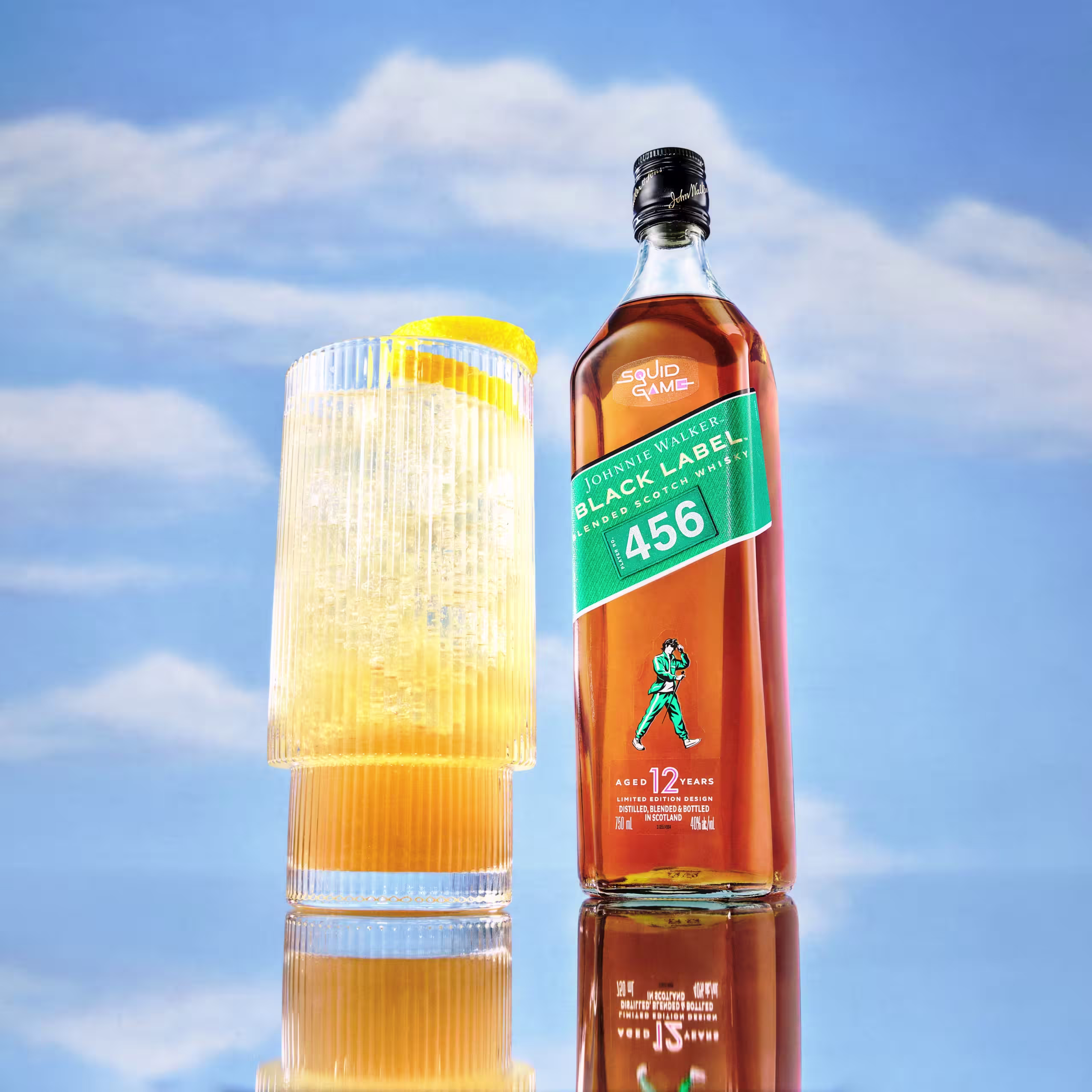 Láhev Johnnie Walker Black Label inspirovaná hrou Squid se zeleným štítkem s číslem 456 a figurkou podobnou hře. Vedle je vysoká sklenice whisky s ledem a ozdobou z pomerančové kůry, postavená na pozadí zatažené oblohy.