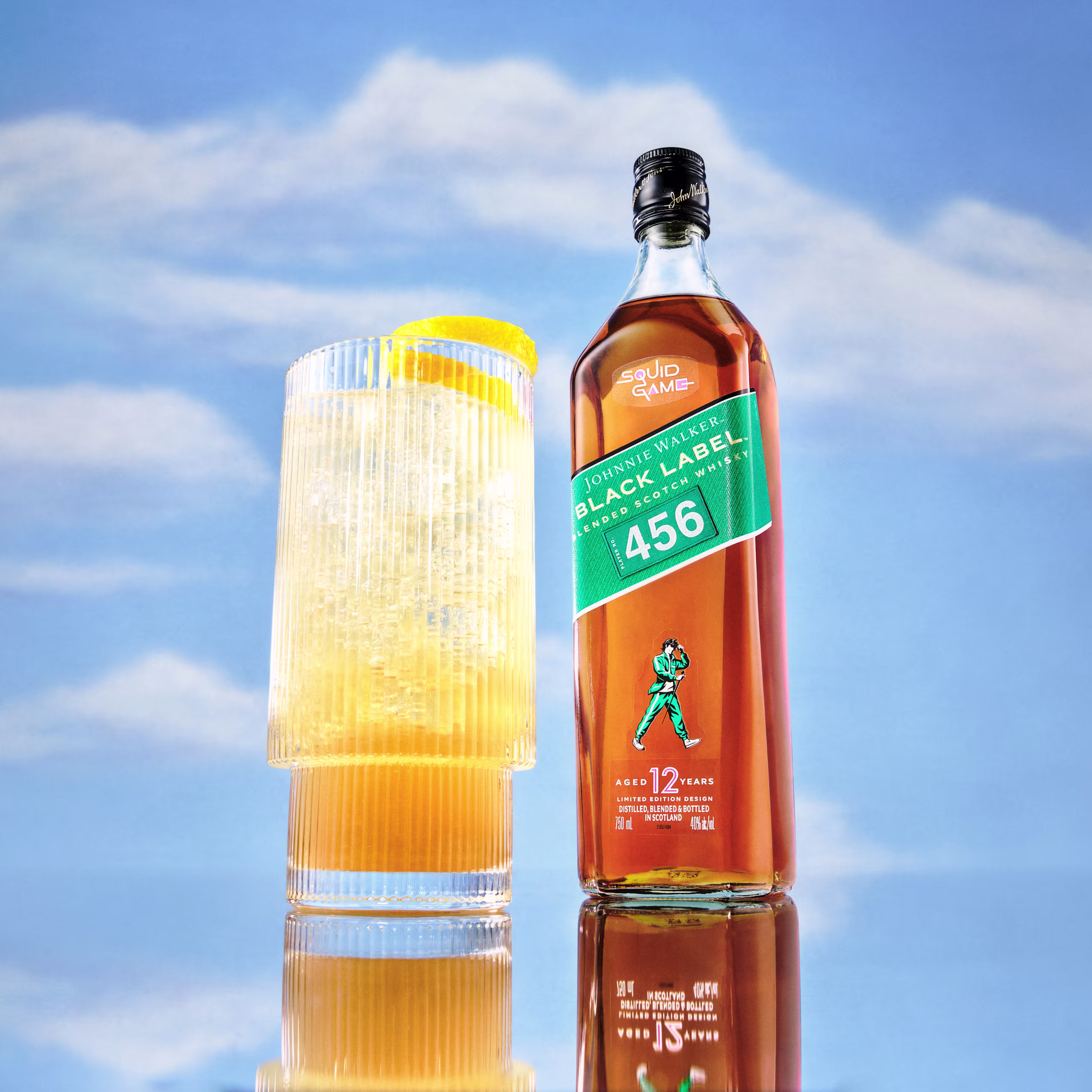 Une bouteille de Johnnie Walker Black Label avec une étiquette « Squid Game 456 » se trouve à côté d'un grand verre de liquide orange avec de la glace et une garniture de tranche de citron, sur fond de ciel bleu avec des nuages doux.