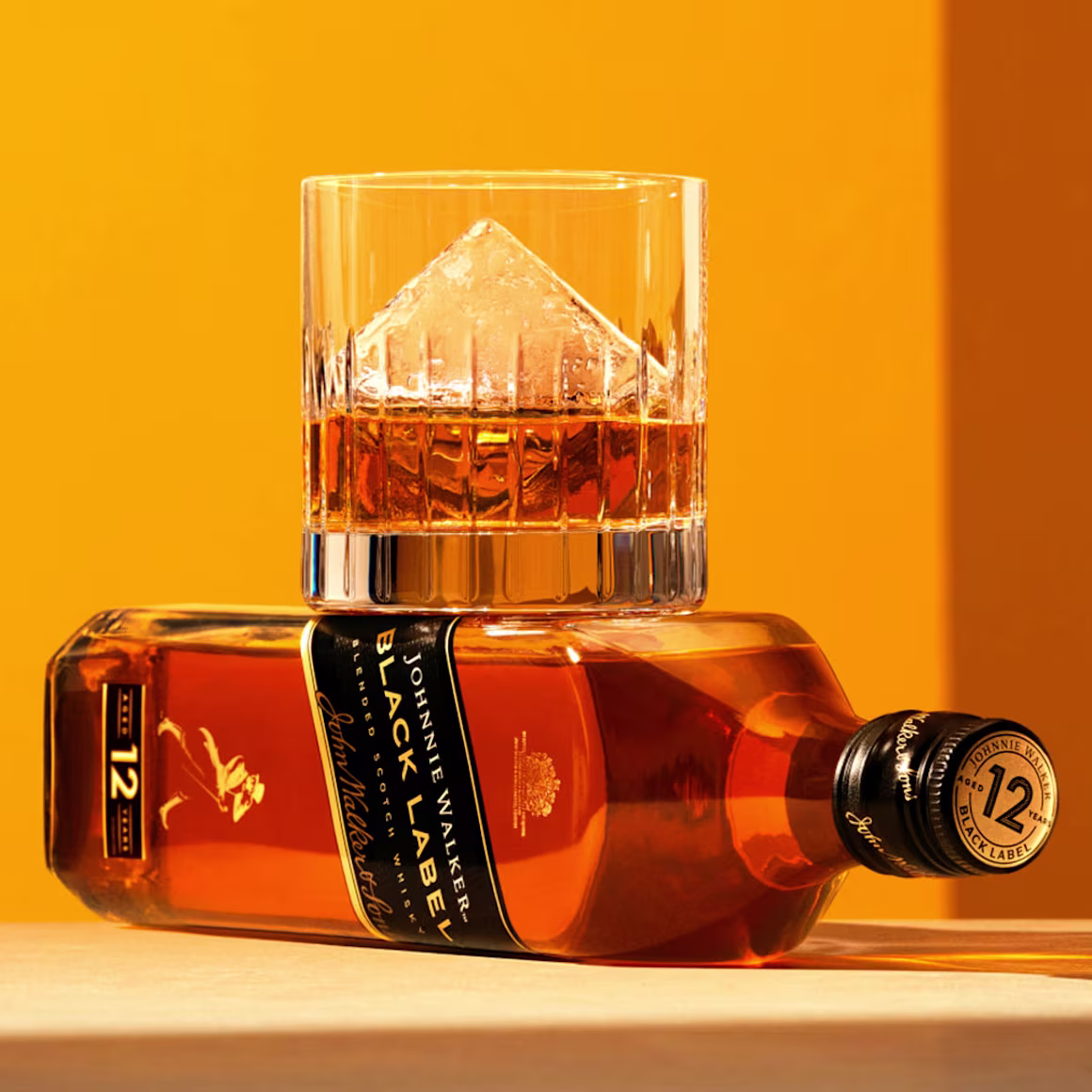 A bottle of Johnnie Walker Black Label whisky lies horizontally on a wooden surface. A glass sits atop the bottle, filled with whisky and a large, pyramid-shaped ice cube. The background is a warm orange color, enhancing the amber liquid.